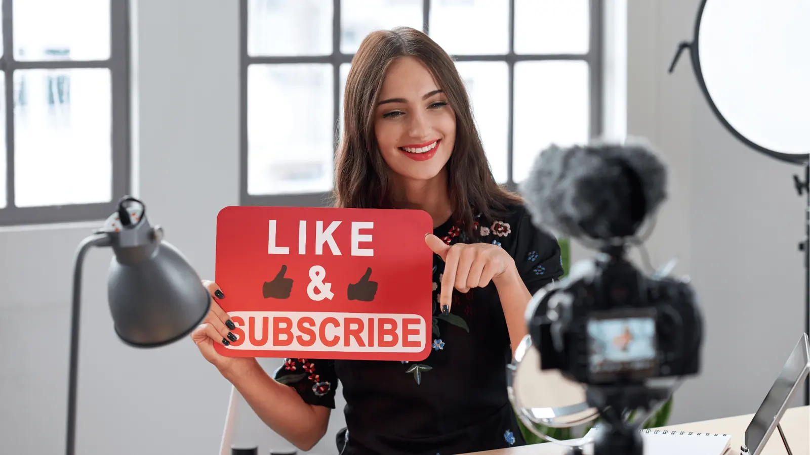 Smiling female content creator holding 'Like & Subscribe' sign in front of camera for AI-assisted workflow