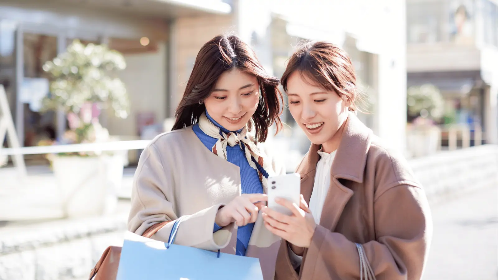 Two women smiling and looking at a smartphone while shopping outdoors