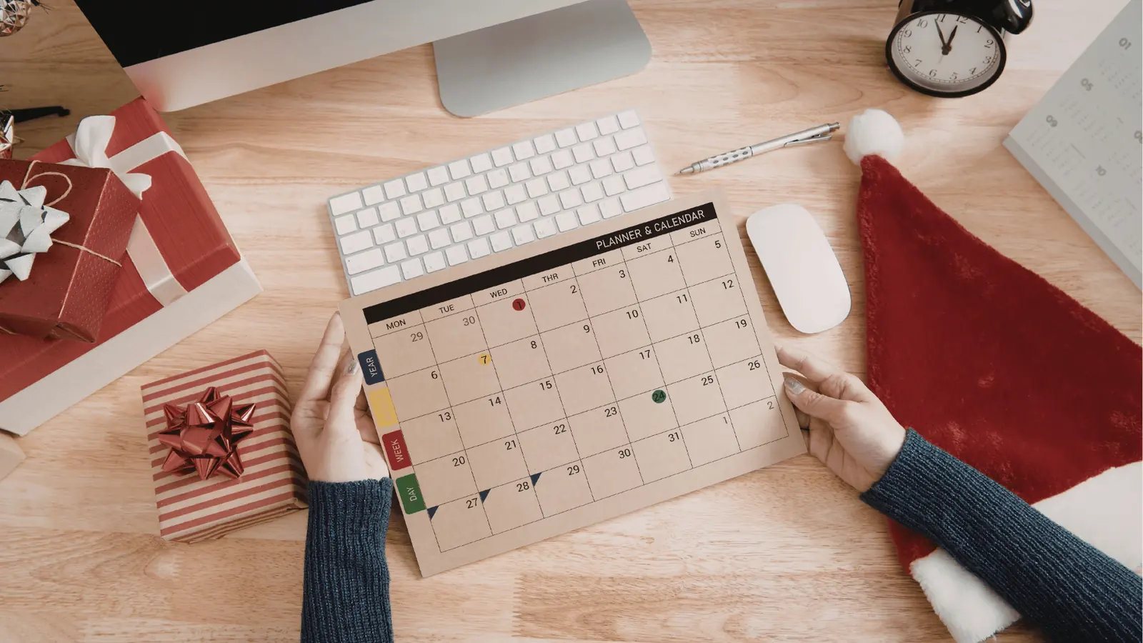 Hands holding a paper monthly planner on a wooden desk with gifts