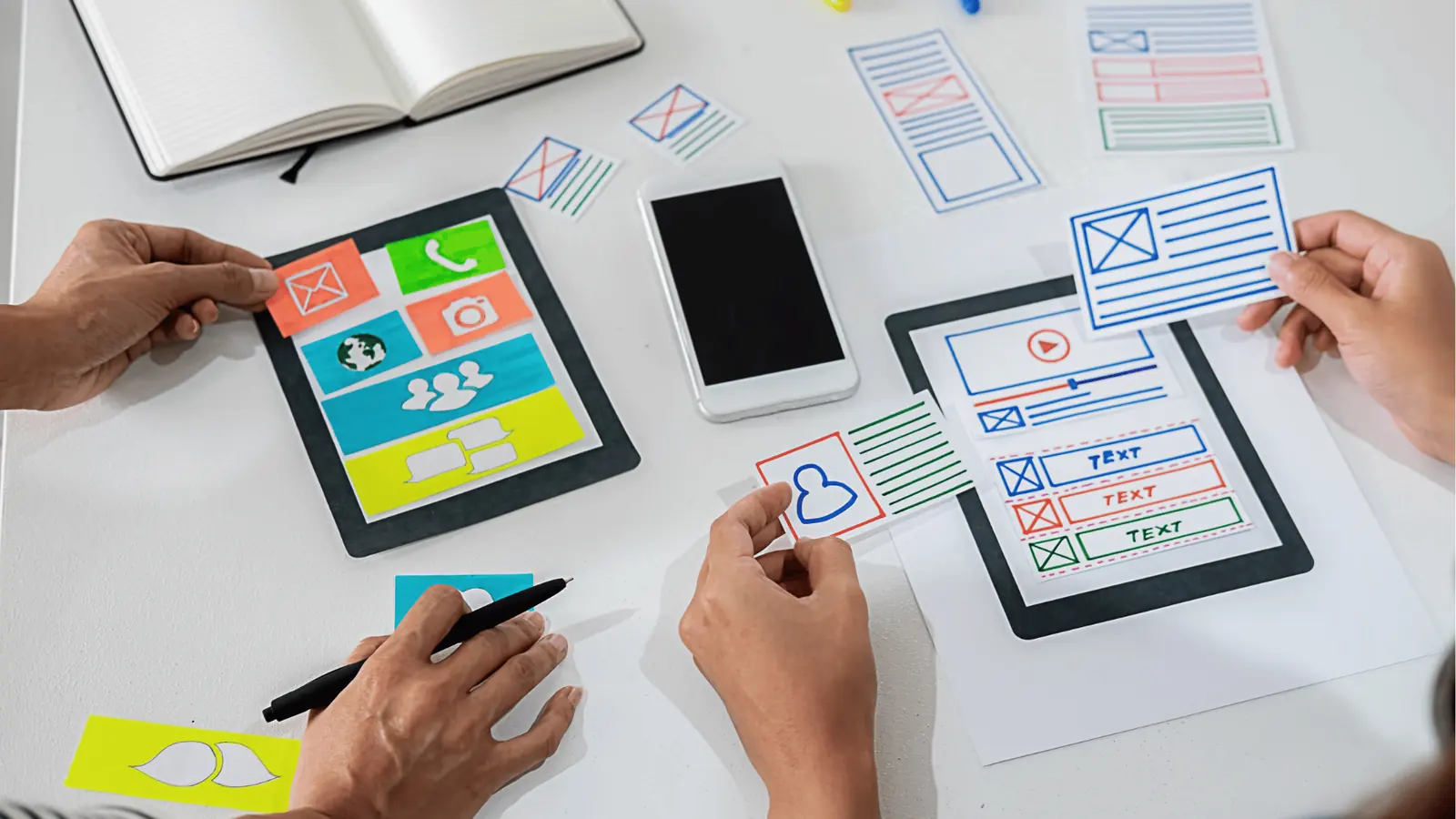 Hands arranging paper mobile app wireframe pieces on a white table for automation
