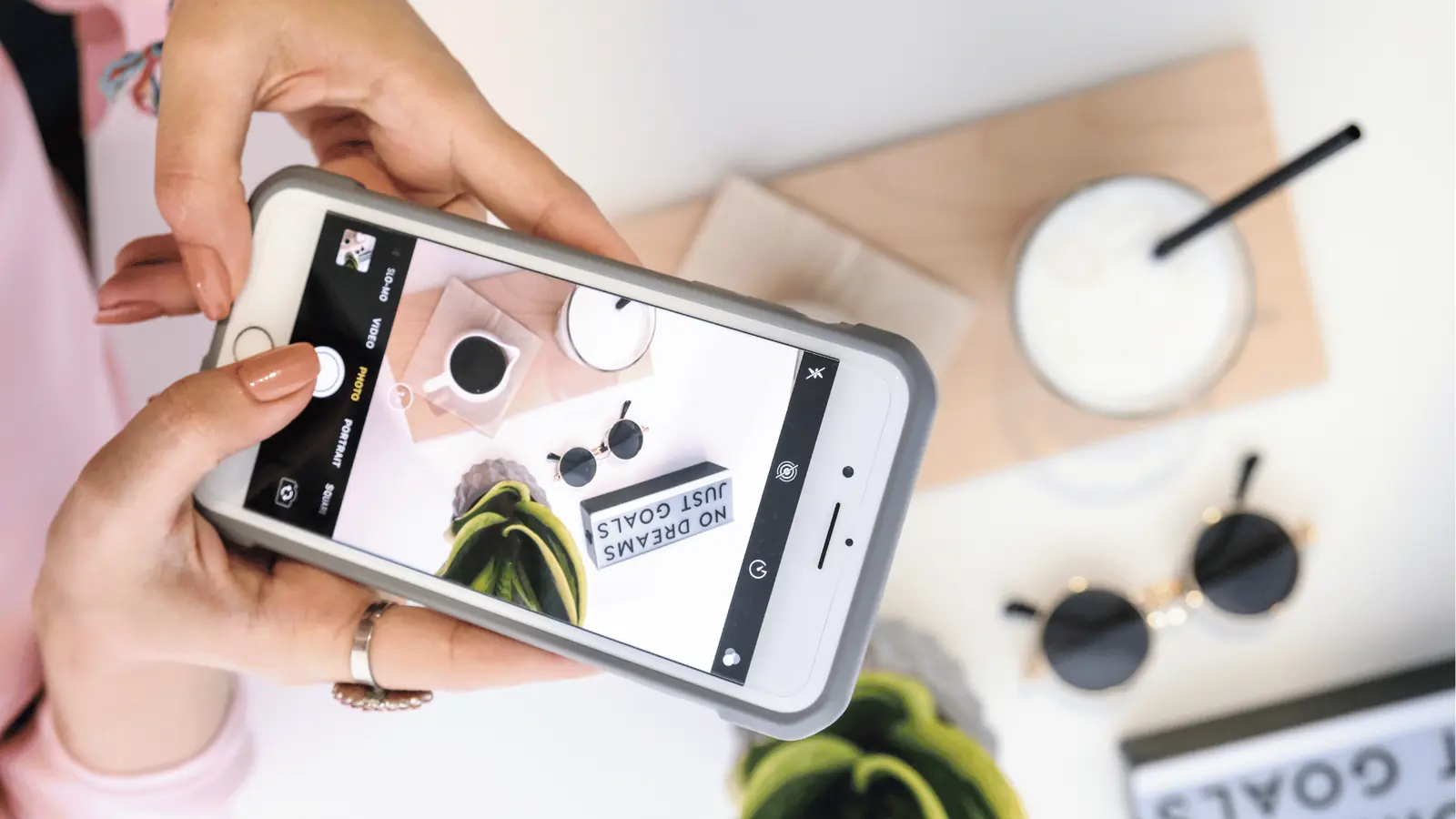 Hands holding phone photographing flatlay with coffee, sunglasses, plant, and inspirational sign