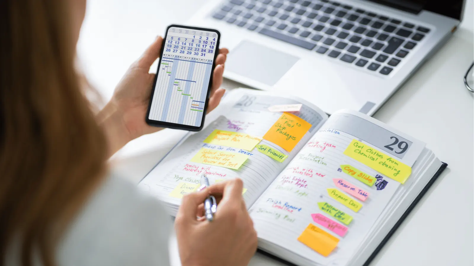 Person holding smartphone showing calendar while writing in sticky-note planner for AI-assisted workflow
