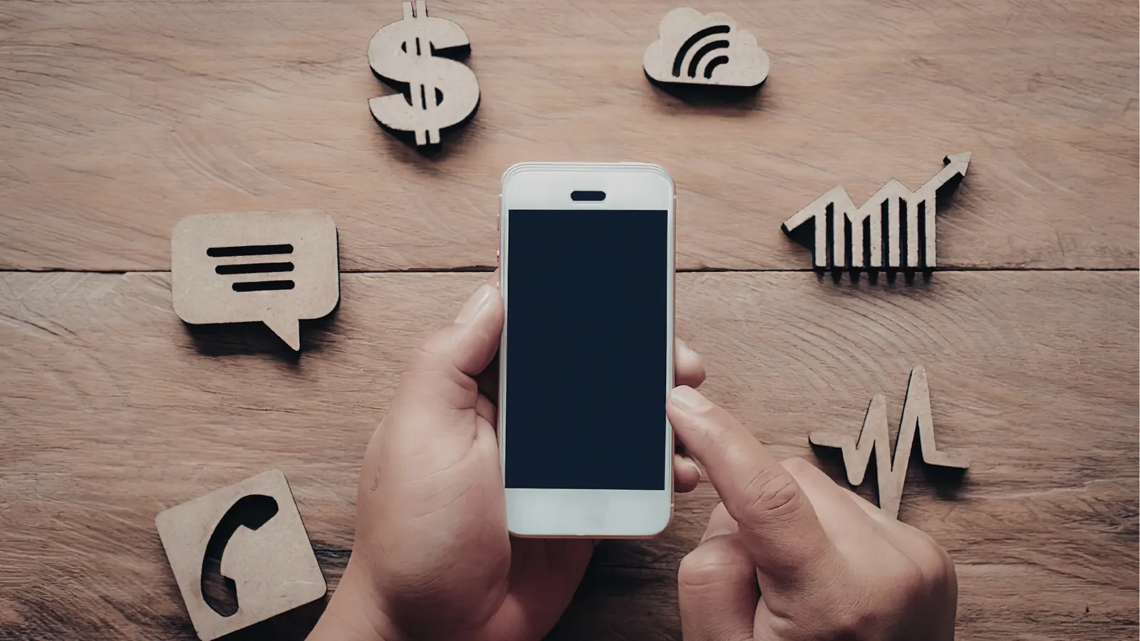 Hand holding smartphone over wooden table surrounded by wooden app icons for publishing