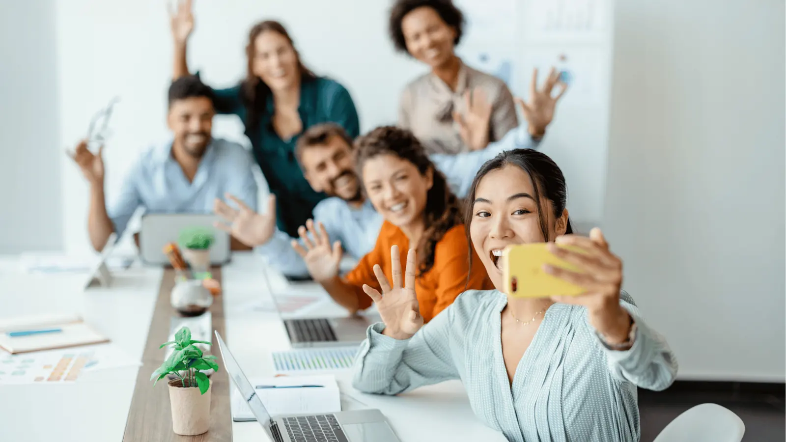 Young woman takes a selfie with smiling coworkers waving at a long office table