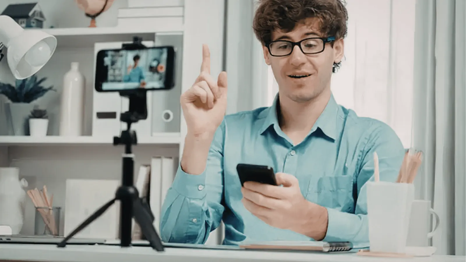 Young man in blue shirt gestures while recording with smartphone on tripod at desk for approval workflow